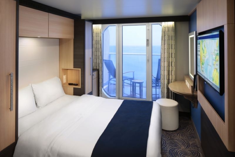 A Studio Ocean View Stateroom with Balcony on board the Anthem of the Seas cruise ship, featuring a cozy bed with white linens and a dark blue runner. The room includes a wall-mounted flat-screen TV, a small desk with a stool, and sliding glass doors leading to a private balcony with seating. The decor is modern and sleek, with light wood tones and a serene ocean view enhancing the relaxing ambiance.