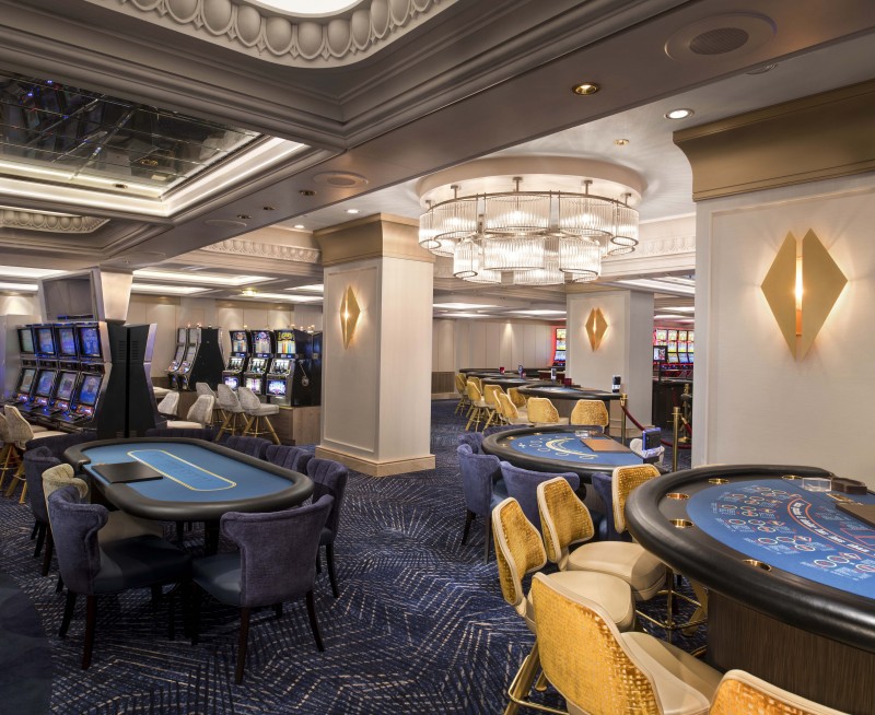 A Celebrity cruises casino's elegant gaming floor with poker tables ready for players, luxurious chairs awaiting guests, and slot machines in the distance, all under soft and sophisticated lighting. The opulent design elements create a welcoming atmosphere for patrons.