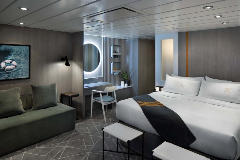 Modern Sky Suite on a Celebrity cruise ship, featuring a cozy bedroom with plush bedding, a stylish sofa, a work desk with a porthole view, and elegant, contemporary decor creating a tranquil retreat for passengers.