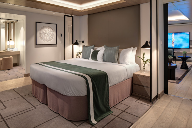 A luxurious Penthouse Suite bedroom on the Celebrity Ascent, elegantly appointed with a large comfortable bed, sophisticated neutral tones, artful decor, and ambient lighting, creating a relaxing and high-end atmosphere for guests.