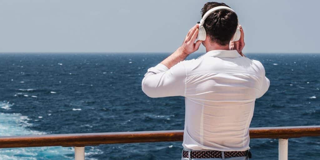 A man stands on a cruise ship deck, gazing out at the ocean, with white headphones on, enjoying the serene view and his music in a moment of relaxation and reflection.