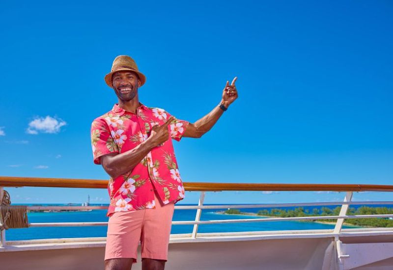 A cheerful guest on a cruise ship, dressed in tropical attire with a straw hat and floral shirt, making a peace sign with the blue sky and sea in the backdrop.