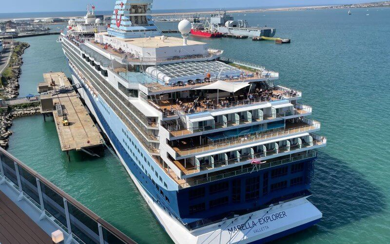 The Marella Explorer cruise ship is docked at a port, showcasing its multiple decks with passengers visible on the open-air top deck, against a backdrop of clear turquoise waters and a distant coastline under a sunny sky.