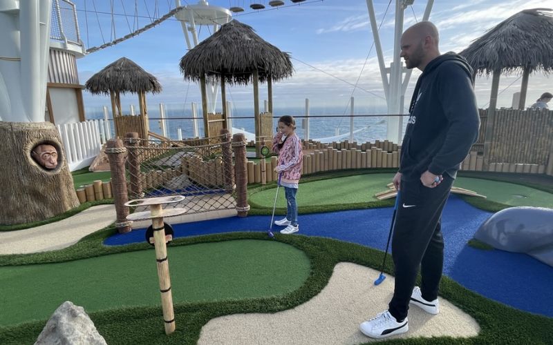 A man and a young girl enjoy a game of mini-golf on a ship deck with a nautical theme, complete with artificial grass, tiki huts, and rope bridges. The ocean horizon extends in the background, adding a serene backdrop to this leisurely activity.