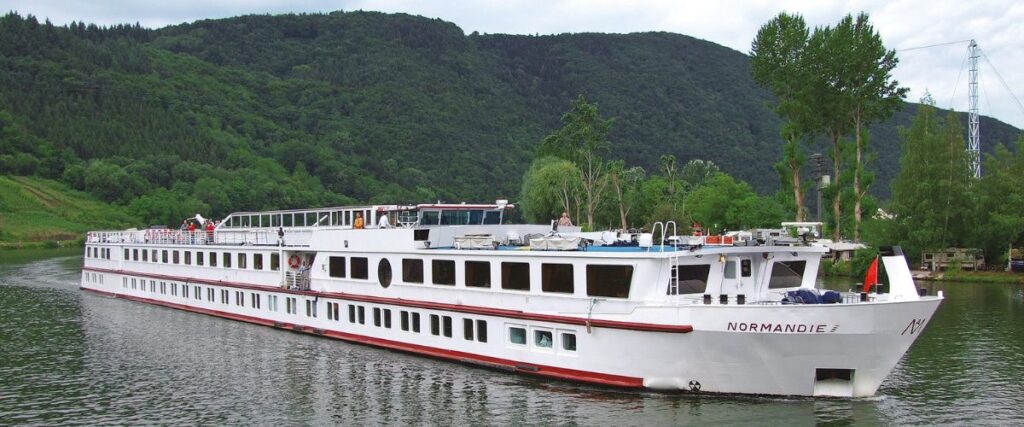 The MS Normandie, a river cruise ship, glides gracefully along a calm river with a backdrop of lush green hills. Its white exterior with red and blue accents reflects a classic and elegant design, providing a leisurely cruising experience through scenic landscapes.