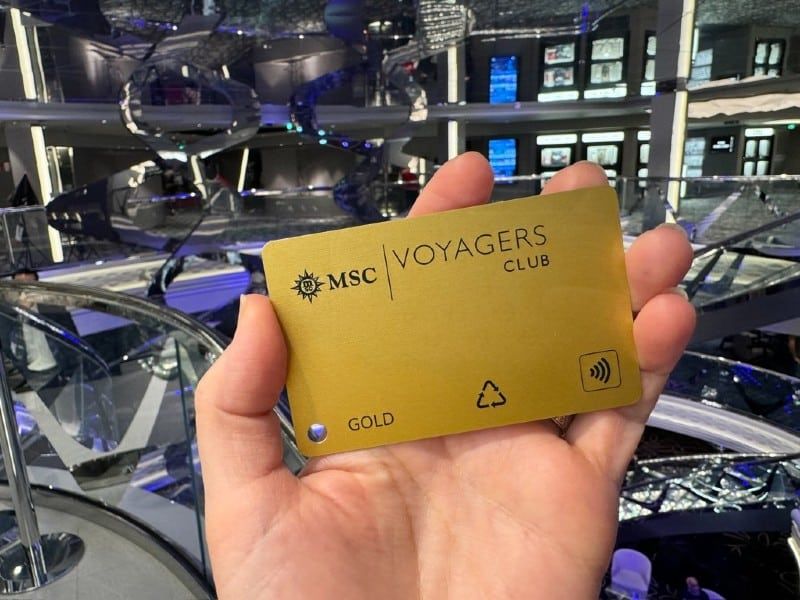 A hand holds up an MSC Voyagers Club Gold membership card, prominently displayed against the backdrop of a cruise ship's opulent multi-level atrium with intricate glass staircases and modern design.