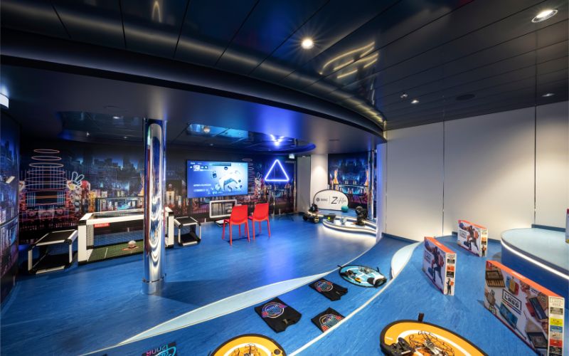 Teens club room on MSC Cruise Line, featuring a futuristic design with blue lighting, gaming consoles, and a large screen for video games. The room includes interactive play areas, red chairs, and shelves displaying various board games and activities, creating an engaging and modern space for teenagers to enjoy.