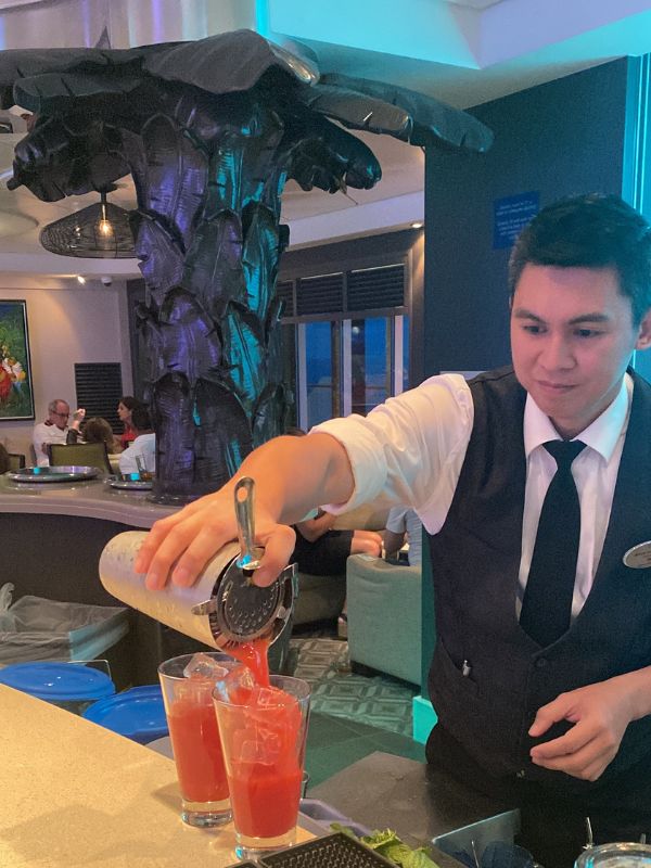 A bartender aboard a Norwegian Cruise Line ship skillfully pours a bright red cocktail into tall glasses, set against the backdrop of an elegant bar with a distinctive modern sculpture and a relaxed atmosphere.