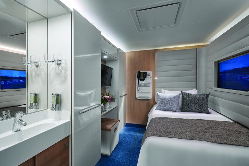 Modern Norwegian Cruise Line Bliss studio room, featuring a cozy single bed with stylish cushions, a wall-mounted TV, a sleek vanity area with a sink and mirror, and a window showcasing a view of the sea.