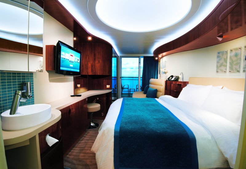 View of a Norwegian Cruise Line Epic stateroom with a balcony, featuring a queen-size bed with white and blue linens, an exposed round basin sink, a wall-mounted TV, and floor-to-ceiling windows leading to the balcony with ocean views.