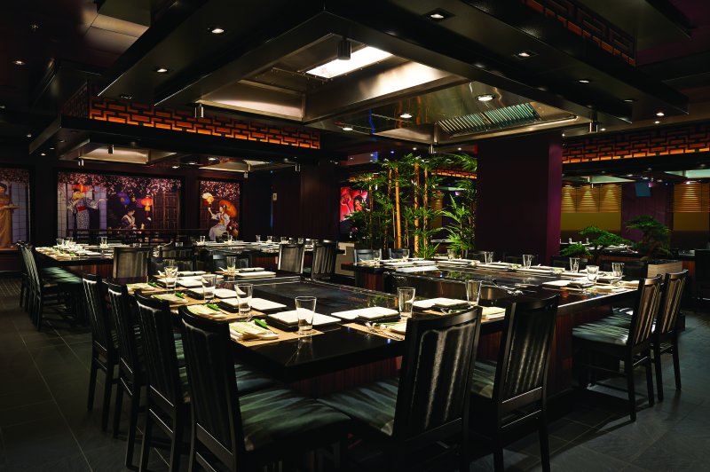 The lively Teppanyaki dining area on NCL Getaway, featuring large communal tables with built-in grills, dark wooden chairs, and vibrant Asian-inspired decor, ready for an interactive culinary experience.