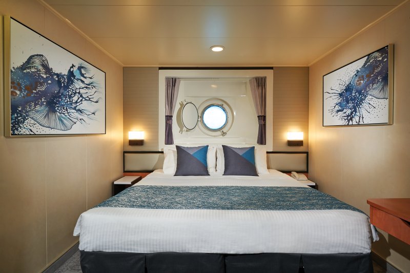 Elegant Oceanview stateroom on the Norwegian Star, showcasing a comfortable bed with a blue bedspread, with porthole window providing natural light, and striking marine-inspired artwork on the walls.