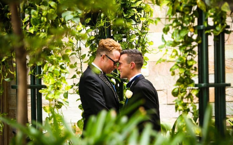 A newlywed couple, dressed in formal suits, share an intimate moment by touching foreheads amidst a lush, green garden. The background features climbing plants and a stone wall, creating a romantic and serene atmosphere.