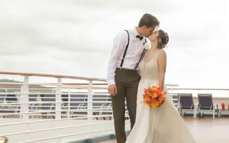 A newlywed couple shares a kiss on the deck of a cruise ship, with the groom wearing suspenders and a bow tie, and the bride in a flowing white gown holding a vibrant bouquet of orange and yellow flowers. The background features a calm, cloudy sky and the boat's railing.