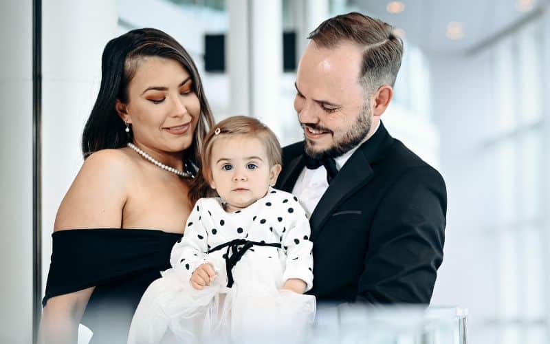 A newlywed couple, dressed elegantly, smiles warmly as they hold their baby dressed in a cute white outfit with black polka dots. The family is positioned in a bright, modern setting, celebrating their special day.