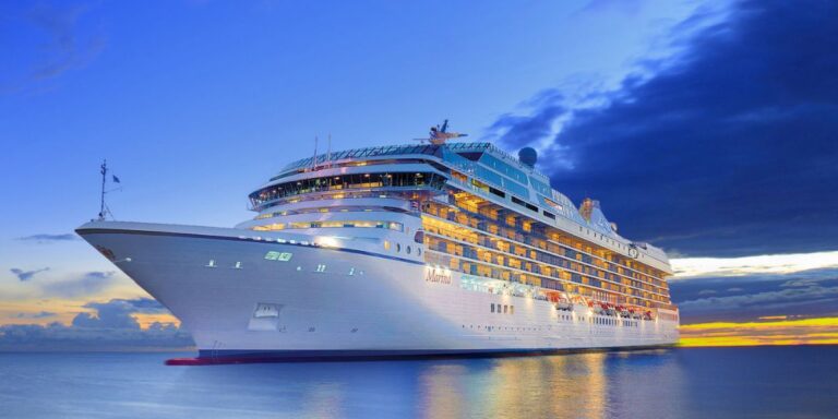 A luxurious cruise ship named "Marina" sailing at sunset with the sky showing vibrant hues of blue and orange. The ship's exterior is illuminated, reflecting the serene water below.