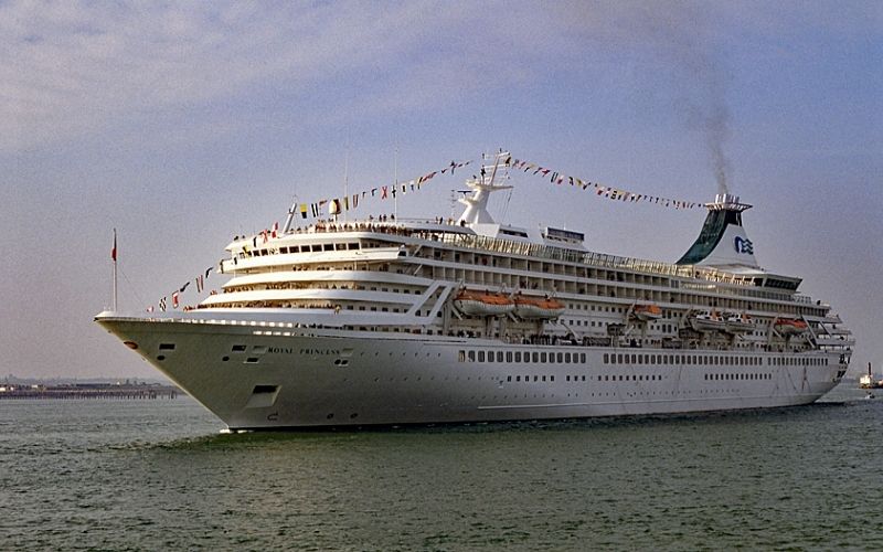 The image shows the classic Royal Princess cruise ship sailing at sea, adorned with festive flags strung along its upper decks. The vessel's white exterior and multiple decks with lifeboats give it a stately appearance, while a gentle plume of smoke rises from its funnel. The scene captures the grandeur of this vintage cruise liner against a calm, blue sky.