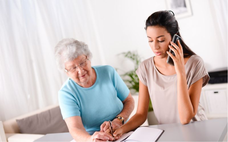 A young woman talks on her phone while assisting an elderly lady in a blue shirt who is writing on a paper, likely involving caregiving or a health consultation.