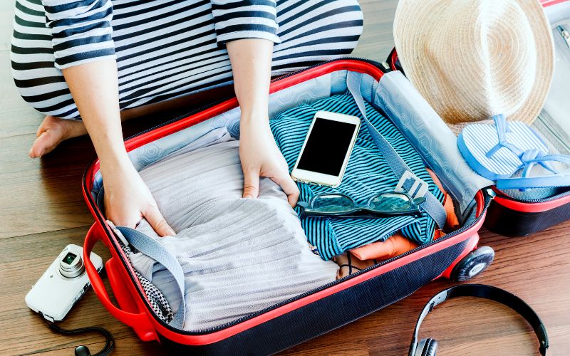 Person in a striped shirt packing a red suitcase with neatly organized clothes, a sun hat, smartphone, camera, and headphones, capturing the process of preparing for a trip with essential travel gadgets and attire.