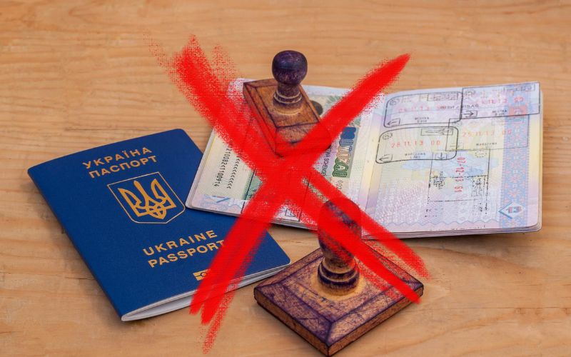 The image features an open Ukrainian passport with visible visa stamps, alongside a wooden stamp, all overlaid with a large red 'X'. This red 'X' could signify a problem or rejection related to travel or visa approval. The setup is on a wooden surface, enhancing the focus on the documents and their significance.