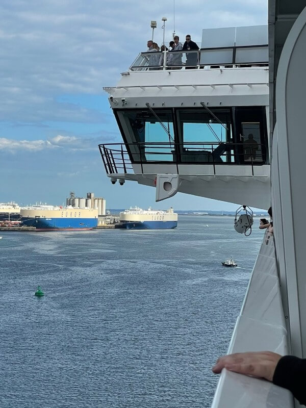 This image captures a unique perspective from a cruise ship's bridge wing, where passengers are seen enjoying the view. The bridge wing extends over the water, offering an unobstructed vantage point. In the distance, a cargo vessel is passing by, contrasted against the industrial backdrop of a port. A small pilot boat moves closer to the foreground, adding to the maritime activity in this scenic ocean view.