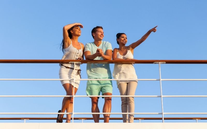 Three friends enjoying a cruise, with a woman pointing towards the horizon while they all stand at the ship's railing against a clear blue sky, capturing the joy and adventure of sea travel.