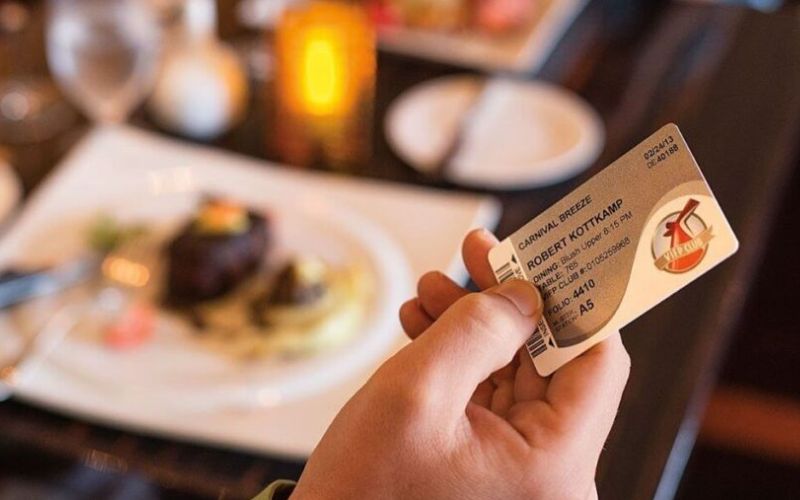 The image captures a close-up of a hand holding a cruise ship boarding pass with the name "Robert Kottkamp" visible, indicating his suite and folio number. In the background, a dining table is set with a gourmet meal, creating an ambiance of luxury and leisure aboard a cruise. The setting underscores the convenience and elegance of cruise dining experiences.