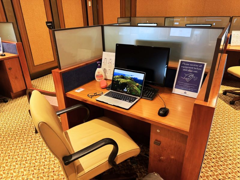 A work area on a Cunard cruise ship, featuring a cubicle with a computer, a laptop, hand sanitizer, a sign reminding guests to sanitize equipment, and a comfortable office chair.