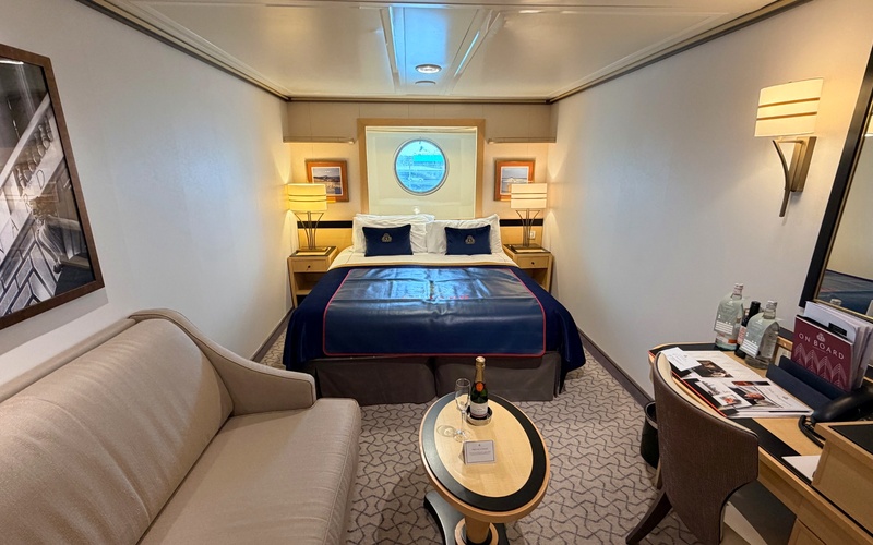 Queen Mary 2 cabin with a double bed beneath a porthole, sofa and small table.