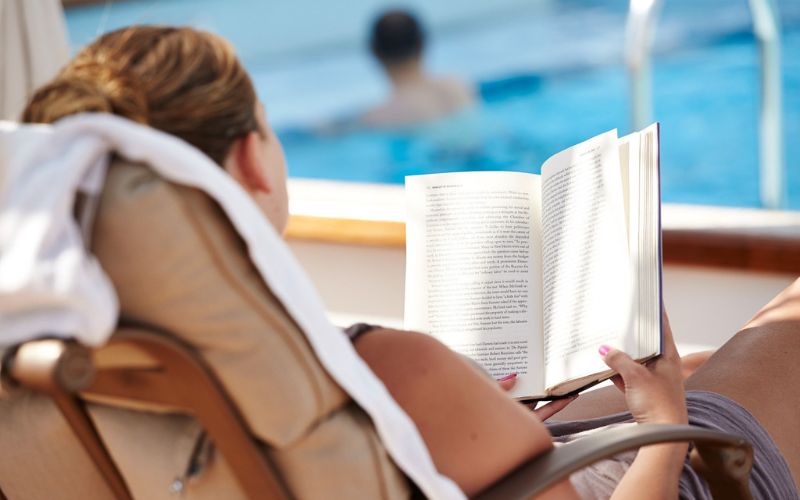 A person is lounging on a deck chair by a pool, reading a book while on a cruise ship. The scene is relaxed, with the pool and another person swimming in the background, creating a serene atmosphere perfect for enjoying a quiet moment with a good book. A towel is draped over the back of the chair, adding to the leisurely vibe.