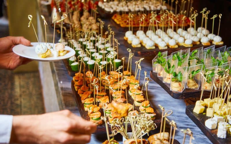 A hand reaches to grab food from a beautifully arranged table of assorted appetizers at a wedding reception, with a variety of bite-sized hors d'oeuvres on skewers and small plates.