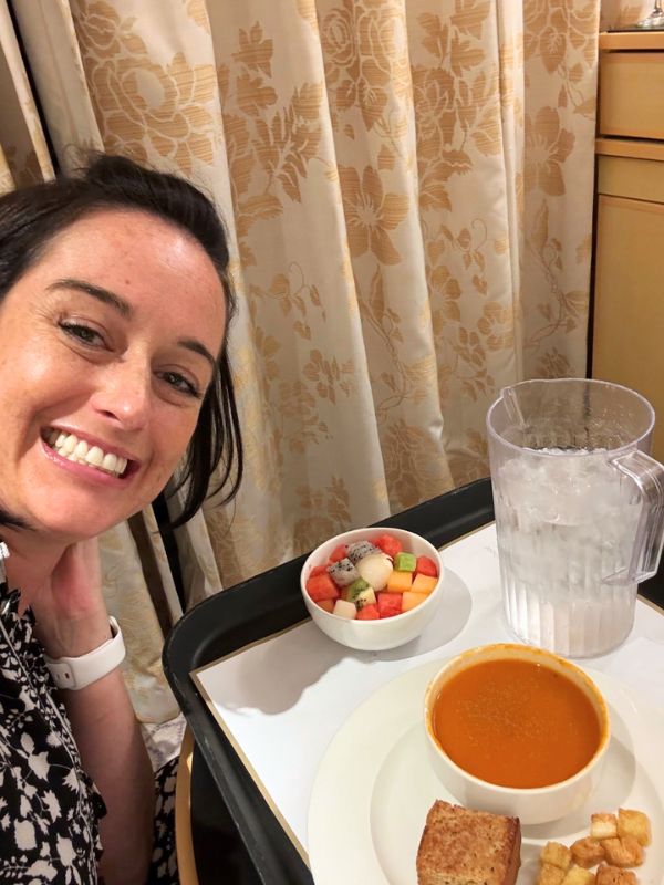 Cruise Mummy smiles beside a room service tray on a Cunard cruise ship, featuring a bowl of tomato soup, a slice of bread, croutons, a bowl of mixed fruit, and a pitcher of water.