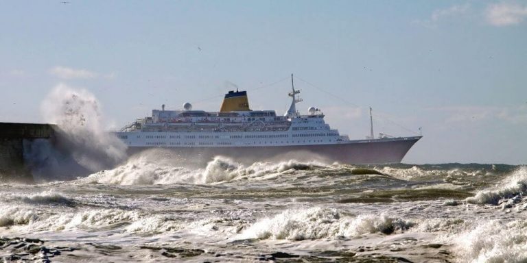 The image captures a cruise ship navigating through rough seas, with large waves crashing against a nearby breakwater and creating dramatic splashes. The ship, framed against a bright sky, appears steady amidst the turbulent waters, emphasizing the power and resilience of the vessel. The scene highlights the contrast between the controlled stability of the cruise ship and the intense, unpredictable force of the ocean.