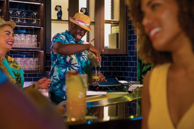 A cheerful bartender in a vibrant blue Hawaiian shirt and straw hat pours a drink at a Royal Caribbean bar, sharing a moment of joy with laughing guests clad in tropical attire, encapsulating the lively spirit of a cruise vacation.