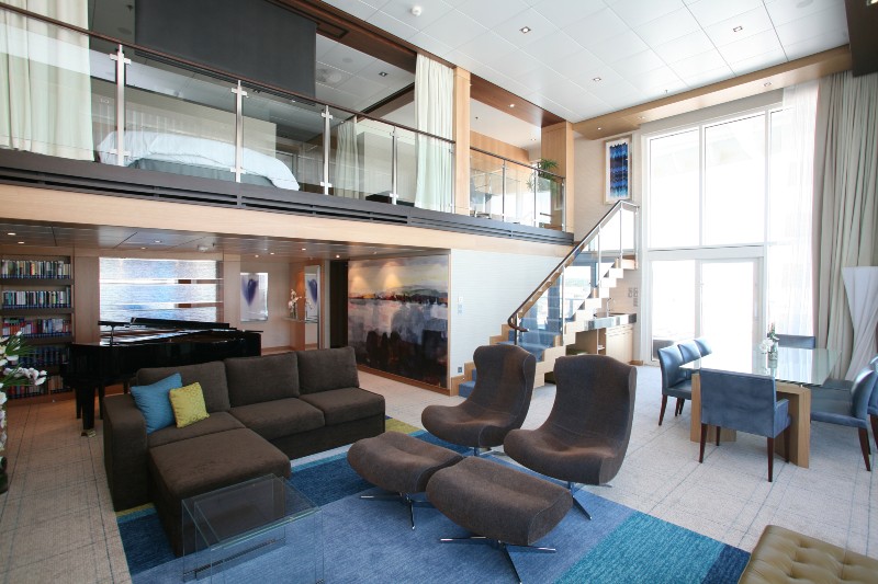 The spacious Royal Loft Suite on the Allure of the Seas, featuring modern furniture, a grand piano, and a two-story window with ocean views.