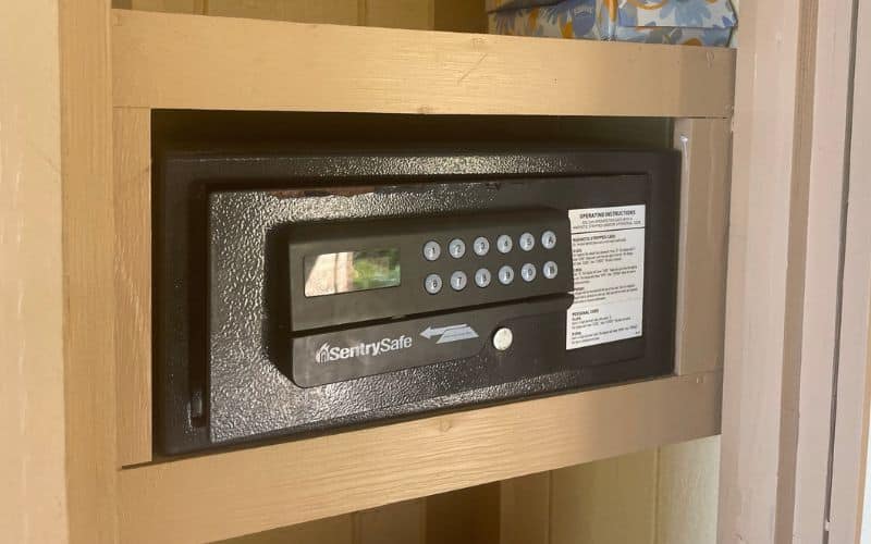 A SentrySafe electronic safe is securely mounted in a wooden compartment inside a cruise ship stateroom. The safe features a keypad for entering a security code and an instructional label on the front. A small decorative box is placed on the shelf above the safe.