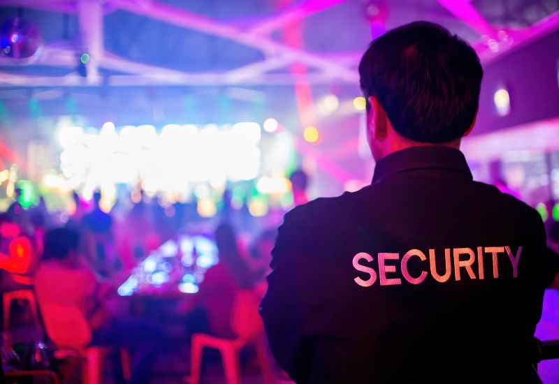 This image shows a security guard on duty at what appears to be a nightclub or event venue aboard a cruise ship. The guard is seen from behind, wearing a black jacket with the word "SECURITY" printed in bold white letters on the back. The background is a blur of colorful lights and a crowd of people, indicating a lively atmosphere with music and dancing. The scene emphasizes the presence of security personnel ensuring safety during onboard events.