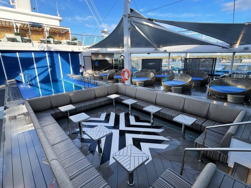 The Serenity Deck on the Carnival Celebration cruise ship, captured on a sunny day featuring a sparkling blue pool, modern grey lounge seating, shaded cabanas, and geometric tables, all inviting relaxation under a canopy sail.