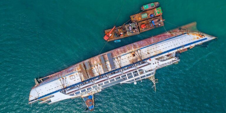 Aerial view of a capsized ship being salvaged at sea, with rust and deterioration visible on its hull, and a salvage barge equipped with a crane moored alongside, highlighting the maritime recovery efforts.