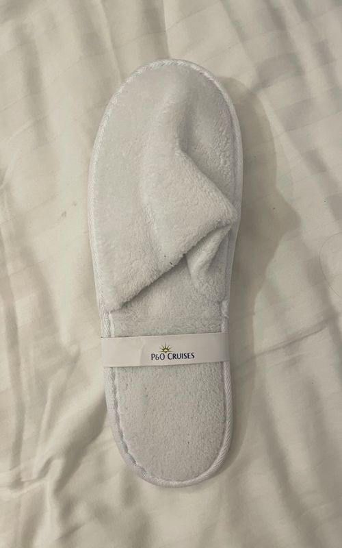 A single plush white spa slipper on a striped bedsheet, with a label around it displaying 'P&O CRUISES', suggesting a comfortable amenity provided on a cruise vacation or a souvenir from the trip.