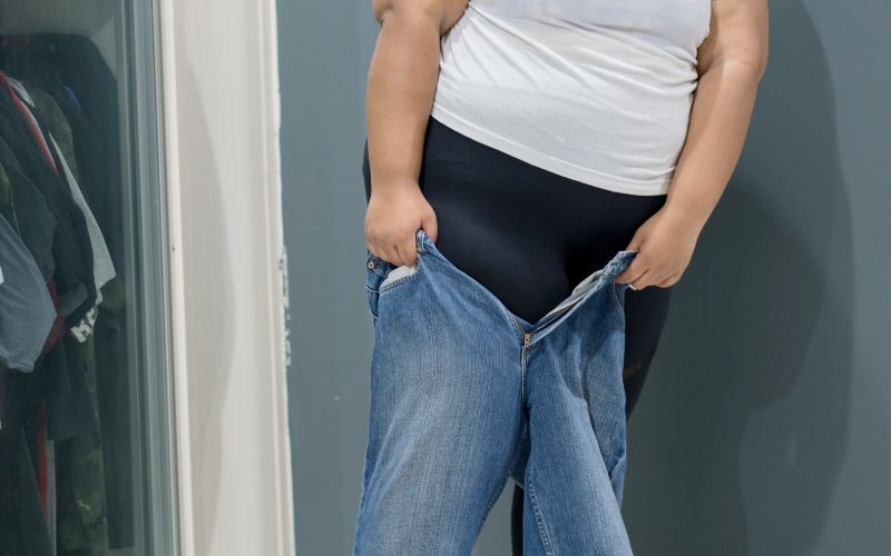 This image shows a close-up of a woman trying to pull on a pair of jeans that are too small. She is wearing a white t-shirt and black leggings under the jeans, which are only partially pulled up to her thighs, clearly illustrating the challenge of fitting into them. The background is simple and neutral, focusing the viewer's attention on the struggle with the jeans. This scene is relatable for many and can be associated with experiences of changing body sizes or the common issue of finding clothes that fit comfortably.