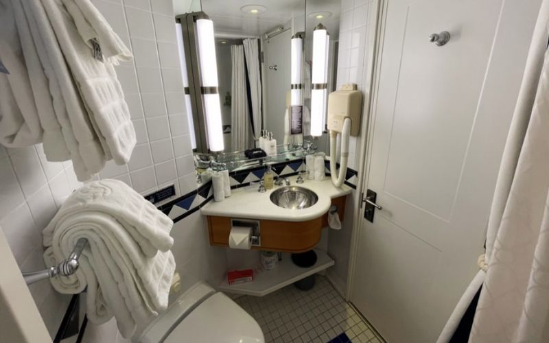 This image depicts a compact and neatly organized bathroom, commonly found in hotel rooms or cruise cabins. It features a white color scheme with blue tile accents. The layout includes a small sink with a vanity mirror, a toilet, and a shower area not fully visible in the image. Towels are neatly hung, and various toiletries are arranged around the sink, creating a clean and functional space for guests.