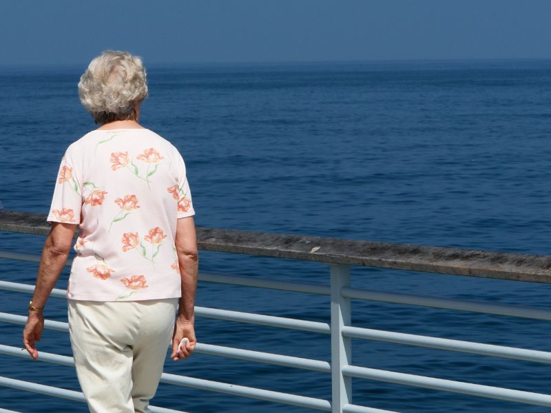 An elderly woman in a floral shirt looking out at the sea from the deck of a cruise ship, contemplating the vast ocean.