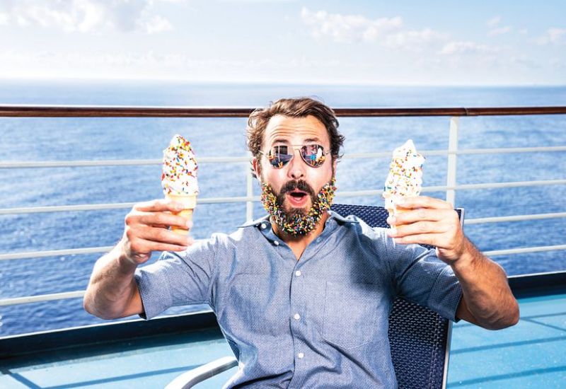 A man with a colorful beaded beard, expressing surprise and delight, holding two ice cream cones on the deck of a cruise ship with the ocean in the background.