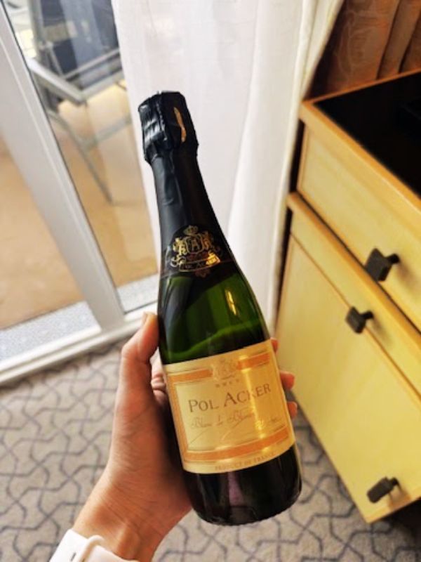 A hand holding a bottle of Pol Acker sparkling wine inside a stateroom on a Cunard cruise ship, with a glimpse of the balcony and outdoor furniture in the background.