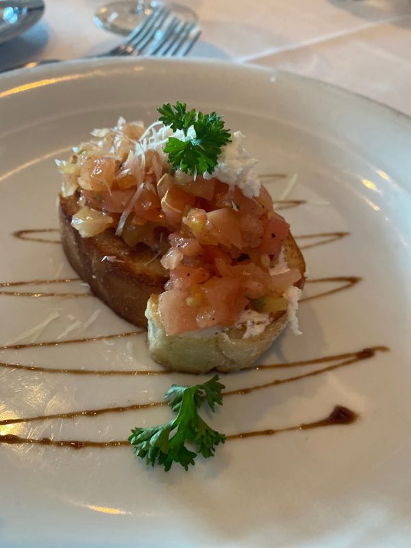 An elegantly plated bruschetta with diced tomato, onion, and a sprinkle of cheese on a toasted baguette slice, garnished with parsley and drizzled with a balsamic glaze, served as part of NCL's specialty dining experience.