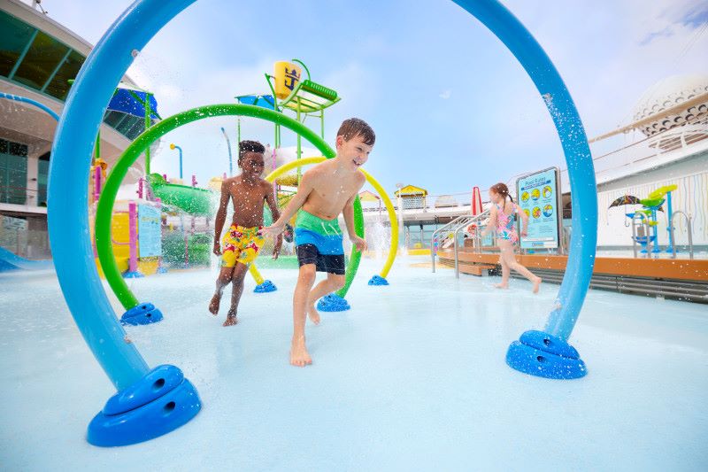 Children splash and play in the sun-drenched Splashaway Bay on Royal Caribbean's Freedom of the Seas. Joyful expressions light their faces as they run through water arches, with a vibrant aquatic playground behind them, capturing the essence of carefree fun on a family-friendly cruise vacation.