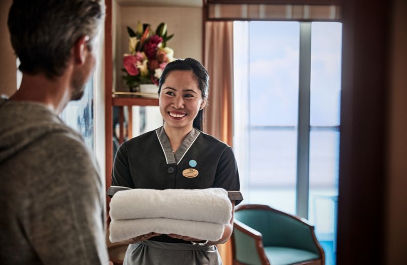 This image shows a friendly interaction between a cruise ship staff member and a passenger in a stateroom. The female staff member, wearing a dark uniform, is smiling and offering a fresh white towel to a male passenger. The background reveals a cabin with a floral arrangement and a view of the ocean through a balcony door.