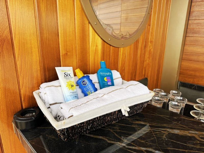 A thoughtful arrangement of sun protection products on the Seabourn Venture, ensuring guests are well-prepared for sunny days during their cruise. Displayed in a wicker basket on a marble countertop, the setup includes bottles of sunscreen with varying SPF levels, an after-sun aloe gel, and fresh white towels for added convenience. This practical setup is placed near a wooden wall with a porthole, emphasizing the ship's nautical theme and attention to guest comfort and care.