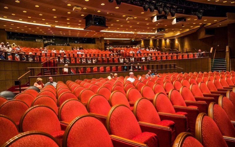 The image displays the interior of a modern theater, with rows of plush red seats facing a stage, not visible in the photo. The theater has multiple levels of seating, including a balcony, all under soft, ambient lighting. The audience is sparsely populated, suggesting either an event before it begins or a moment of calm before the performance resumes. The layout and design emphasize comfort and an immersive viewing experience for the attendees.